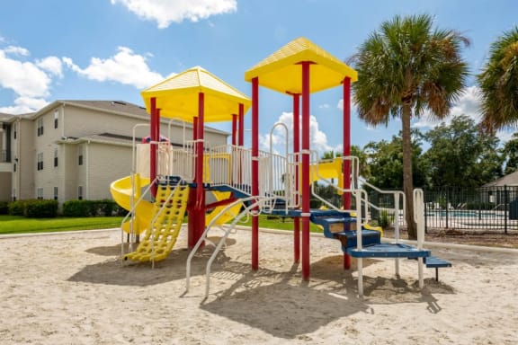 Blue Red and Yellow Playground on Sand with Building Exterior and Treeline in the Background