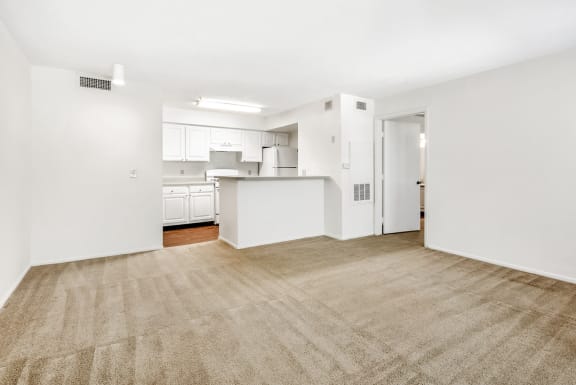 Vacant living room with wall-to-wall carpet. Kitchen in the background with white cabinets and appliances.