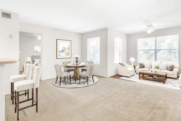 Staged living room with a dining room with hardwood-style flooring and carpet combo, a ceiling fan, and several windows with window blinds.
