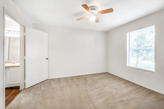 A vacant bedroom with carpet, a ceiling fan, window, and an en suite bathroom.