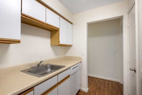 Vacant galley kitchen featuring hardwood-style flooring, white cabinets with wood trim, and a white dishwasher sitting adjacent to the kitchen pantry. Stainless steel sink sits atop beige countertop. The far end of the kitchen opens up to the bedroom hallway.