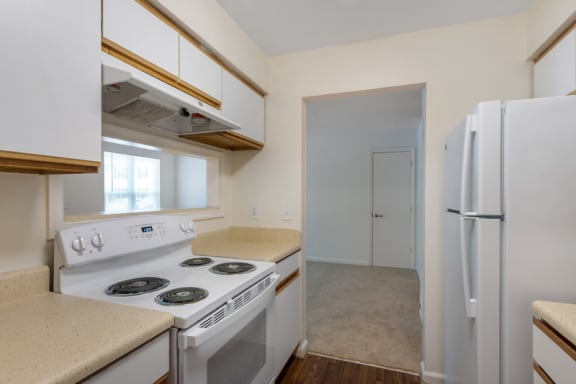 A vacant three-bedroom kitchen featuring wood-style flooring, white cabinets with wood trim, and white appliances including oven, stove, hood vent, and refrigerator. The kitchen opens up to the dining room via a bar-top cutout.