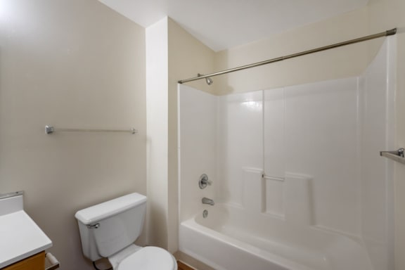 A vacant bathroom featuring off-white walls and a white ceiling. Brown wood-style flooring throughout. The bathroom also features a white shower/tub combo along with white bathroom vanity, white sink, and wood-colored trim. A large frameless bathroom mirror sits above the vanity  A Silver shower rod is in place for a curtain. A white toilet rests in between the vanity and tub.