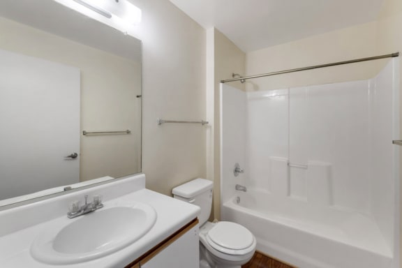 A vacant bathroom featuring off-white walls and a white ceiling. Brown wood-style flooring throughout. The bathroom also features a white shower/tub combo along with white bathroom vanity, white sink, and wood-colored trim. A large frameless bathroom mirror sits above the vanity  A Silver shower rod is in place for a curtain. A white toilet rests in between the vanity and tub.