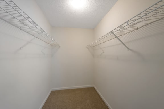 A vacant walk-in closet with carpet throughout, white walls, and white wired shelving installed on either side of the closet.