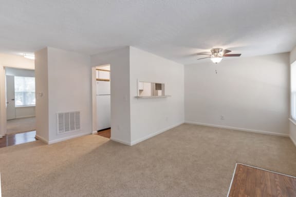 A vacant three-bedroom living room featuring white walls and ceilings. The vinyl floor at the entry gives way to a carpeted living room area and dining room area. There is a small cut-out with breakfast bar opening from the kitchen to the dining room. The kitchen features hardwood style flooring with white appliances and the second bedroom is located in the background.