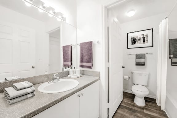 Virtually staged bathroom with large mirror, wood plank flooring, gray countertop, toilet and tub with white shower curtain