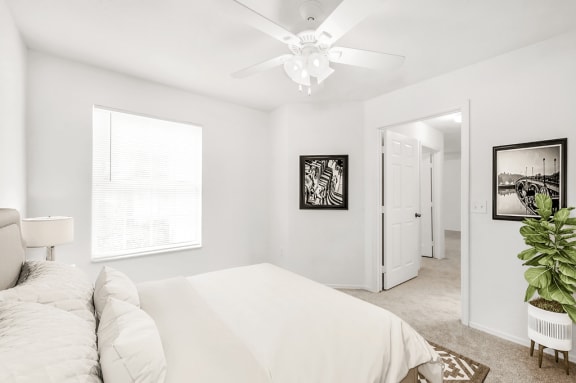 Virtually staged bedroom with carpet, white walls, ceiling fan with light, large window, accent rug, and plant decor.