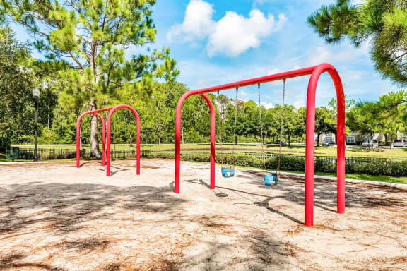 Two red swing sets in the playground area surrounded by an open grass area and trees.