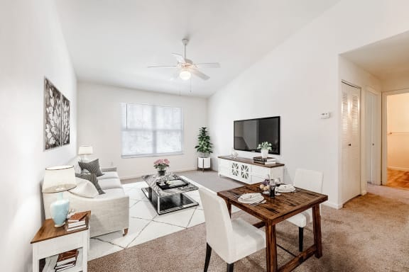 Staged living room with a dining room with carpeted flooring, a ceiling fan, mounted tv, and a windows with window blinds.