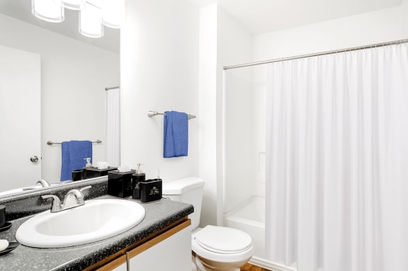 The bathroom also features a white shower/tub combo along with a black granite style bathroom vanity, white sink, and wood-colored trim surrounding the white cabinets