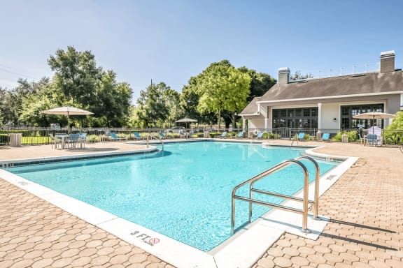 Swimming pool with large tanning deck, blue chairs, table with umbrella, clubhouse in background and surround native landscape