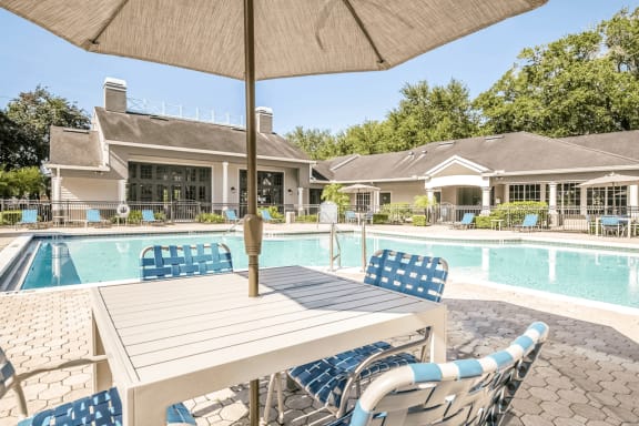 Swimming pool with large tanning deck, blue chairs, table with umbrella, clubhouse in background and surround native landscape