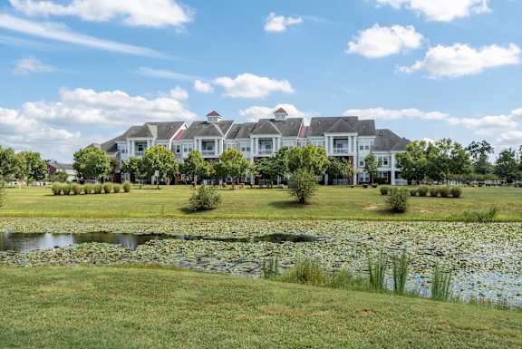 A large white house with a red roof is surrounded by a grassy lawn and a pond with lily pads.