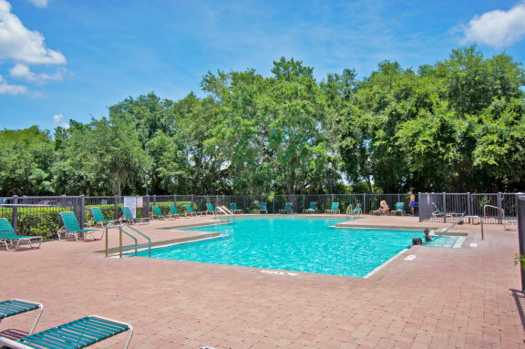 Community pool with sundeck, lounge chairs, surrounded by grey fence with trees, in the background
