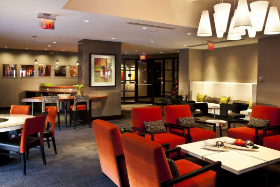 Clubroom seating area. Multiple orange accent chairs with white coffee table. Kitchen in background with bar seating and additional seating behind bar with black accent chairs.