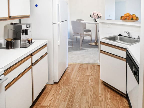 Vacant apartment kitchen with hardwood-style flooring, white wood trimmed cabinets, white appliances and countertops. Virtually staged with a coffee machine, a bowl of oranges on the bar top and a dining table with four chairs in the background.