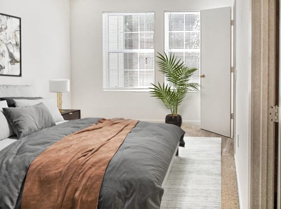 Bedroom with two windows, white walls and beige carpet virtually staged with a bed, wall art, side table with lamp and a standing plant