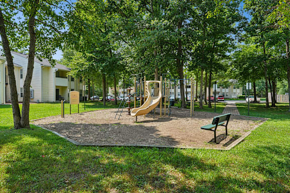 A playground with a slide, swings, and a bench is surrounded by trees.
