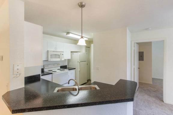 Fully-Equipped Kitchen with Double Basin Sink with Breakfast Bar Black Counters White Cabinets and White Appliances and Doorway to Carpeted Bedroom