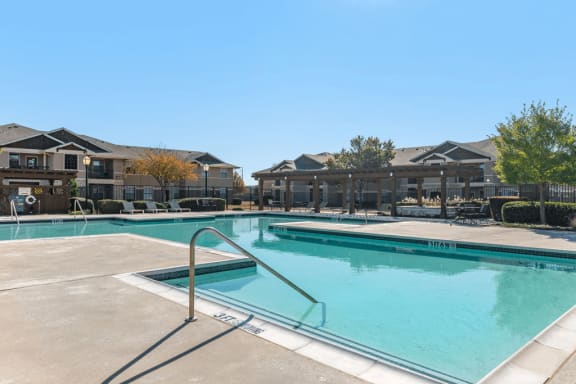 Swimming pool with large tanning deck, lounge chairs, pergola, building exterior in background and native landscape