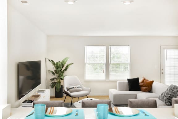 Virtually staged carpeted living room with large window next to entry door. Grey couch and accent chairs with tv in background
