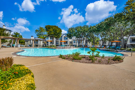 A large swimming pool surrounded by a sandy area and green plants.