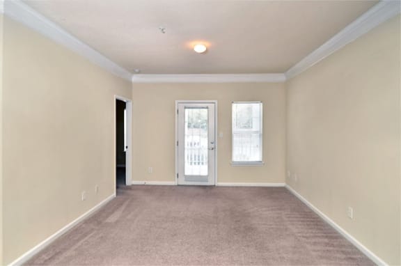 Vacant apartment living room with tan carpet and beige walls. The patio door is on the back wall of the room.