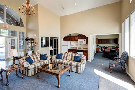 Clubhouse Seating Area with Large Windows Striped Couches with Throw Pillows Next to Coffee Table and End Table with Clubhouse Kitchen in The Background with Wood Cabinets and White Appliances and Business Center Table Further in The Background