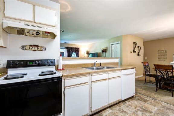 Model Kitchen with White Cabinets and Appliances Beige Counters with Dining Room Table Visible in the Background
