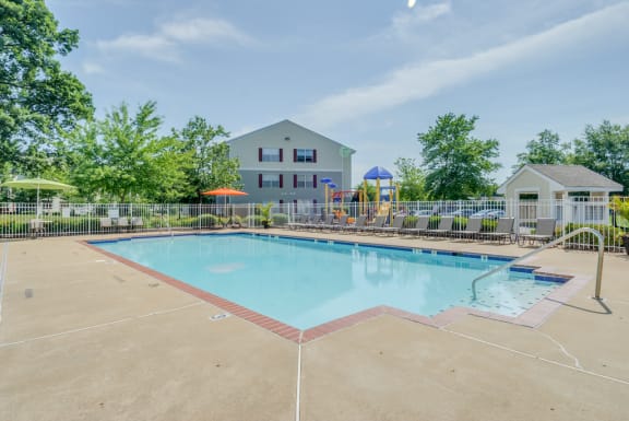 The community pool is rectangular in shape and is surrounded by a spacious sundeck with taupe pool lounge chairs, and two round picnic tables with umbrellas in the middle of the table. The pool is enclosed by a white iron fence. Outside of the pool area is the community playground, the mailbox station, apartment buildings, and mature trees.