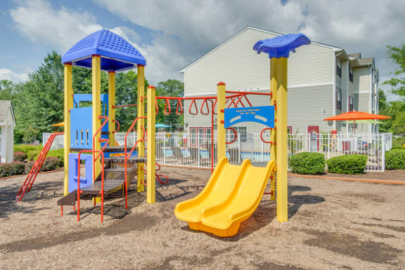 The community playground is red, yellow and blue. It has a ladder, monkey bars, stairs, and two yellow slides. The playground area has mulch on the ground and is located near apartment buildings and the community pool.