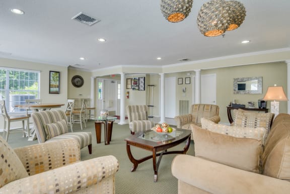 The community clubhouse has four striped chairs, a large tan couch, two glass-topped tables, a coffee table, a round high-top table with coordinating chairs, a large picture window, and three white columns located along the walkway near the front door. There are various pieces of framed art hanging throughout.