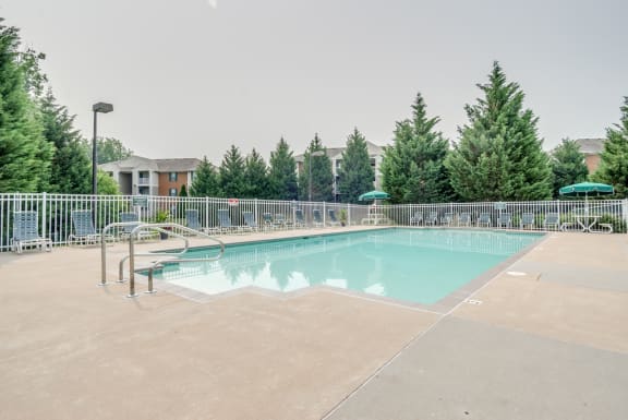 The community pool is rectangular in shape with blue and green pool furniture placed around the sundeck. There is a round picnic table with a green umbrella in the middle. The pool is placed on the back side of the Leasing Office.