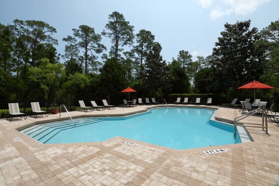 The swimming pool is a large area with calm turquoise waters with a brick paver sundeck surrounding. Light lounge chairs and orange umbrellas are placed around the sundeck.