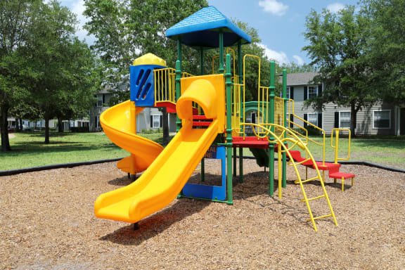 The playground equipment has a yellow slide and yellow metal stairs and poles. There are red steps and a blue roof. The equipment is in a mulched area with a large grassy area surrounding.