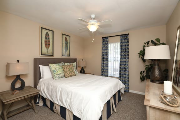 The Bedroom is a comfortable space with a large bed centered on the main wall. A large window brings plenty of natural light into the room. The carpeted flooring continues throughout with furniture filling spaces around the room.
