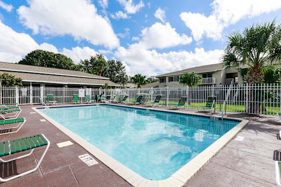 Community pool with sundeck, lounge chairs, with lake view surrounded by white metal fence with palm trees and building exteriors, palm trees, and trees in the background