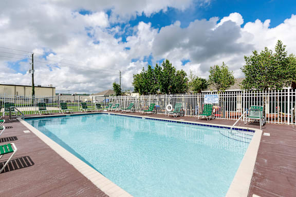 Community pool with sundeck, lounge chairs, with lake view surrounded by white metal fence with palm trees and building exteriors, palm trees, and trees in the background