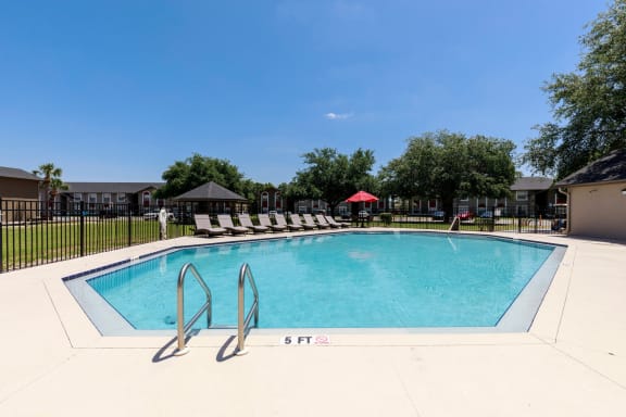 Community pool with sundeck, lounge chairs, with lake view surrounded by white metal fence with palm trees and building exteriors, palm trees, and trees in the background
