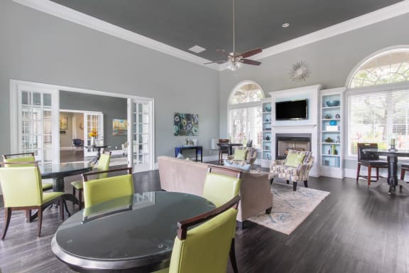 Clubhouse Seating Area with Large Windows on either side of Fireplace with TV and Shelves Surrounding Fireplace Multi Colored Accent Chairs Grey Couch and Dining Room Tables with Green Chairs and Clubhouse Foyer in the Background