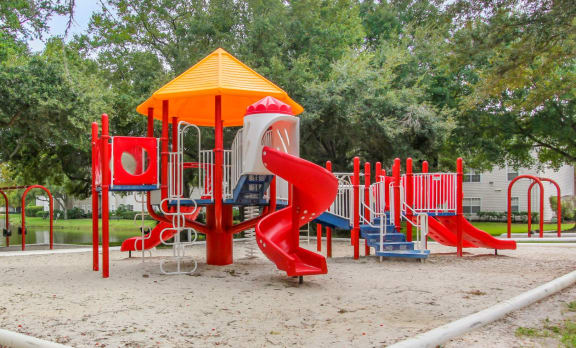 Blue Red and Yellow Playground on Sand with Building Exterior and Treeline in the Background