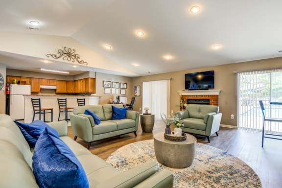 The community clubhouse has tan walls, hardwood style flooring, a fireplace located between two sliding glass doors with vertical blinds, and a kitchen area with white appliances, wooden cabinets and breakfast bar that opens to the remainder of the room.
