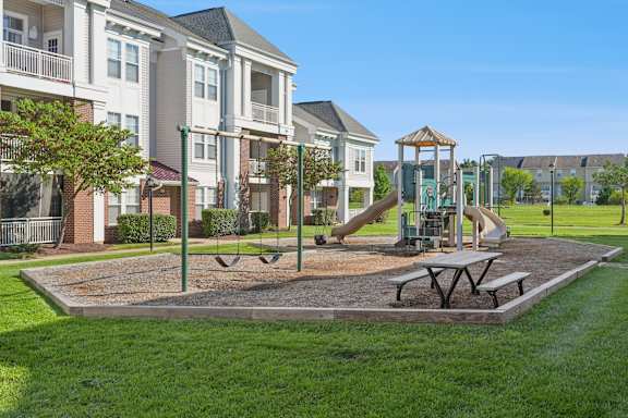 A playground with a swing set and picnic table in front of apartment buildings.