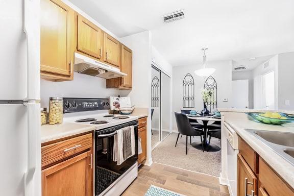 A galley-style kitchen with a double basin sink, wood cabinetry, and a dishwasher across from a white refrigerator and electric stove. A dining area with a mirrored wall and a round dining table with four chairs are in the background.