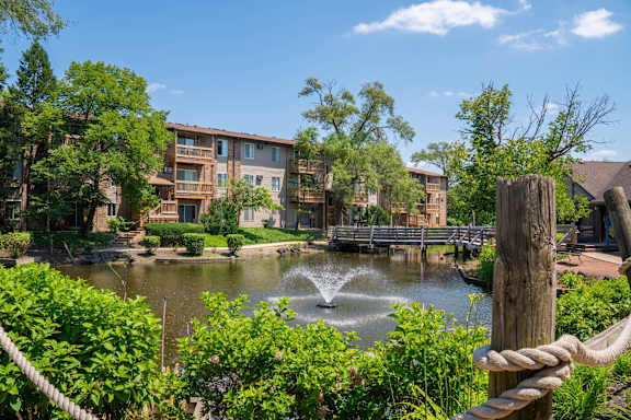 A fountain in the middle of a pond surrounded by greenery and a building in the background.