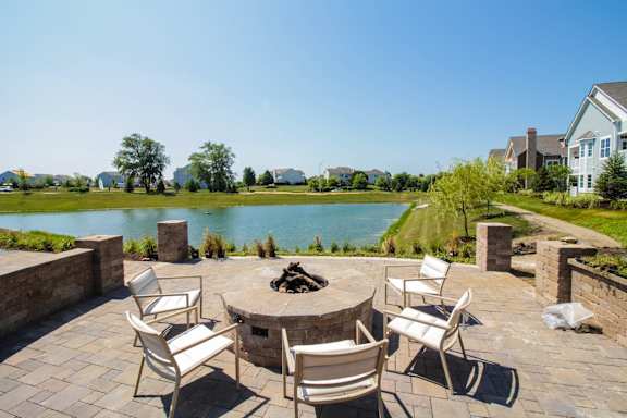 A patio with a fire pit and chairs overlooks a lake.