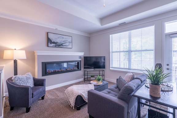 A living room with a grey couch, a grey chair, a grey ottoman, a white blanket, a fireplace, a television, a potted plant, and a painting on the wall.