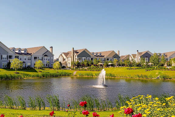 A serene lakeside scene with a fountain and houses in the background.