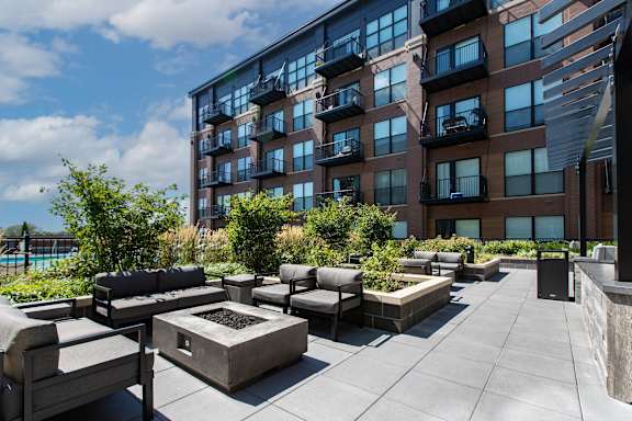 A modern outdoor seating area with a fire pit is surrounded by a large building.